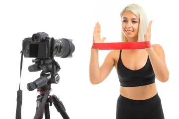 A young sports girl with blonde hair and bright manicure in black sports topics, leggings and sneakers does exercises with sports rubber in front of the camera.