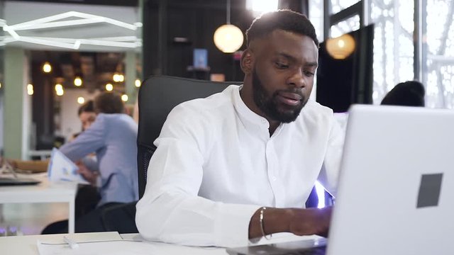 Dissappointed dark-skinned bearded businessman seriously looking at the computer screen at his workplace on the background of office interier with workers