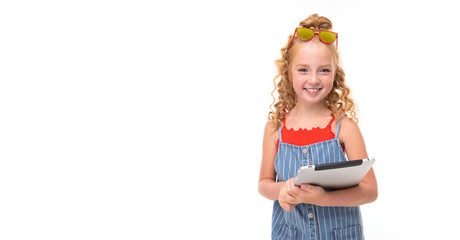 A little girl with red heap hair in a red jersey and blue-and-white jumpsuit in a stripe holds a tablet. and smiling.