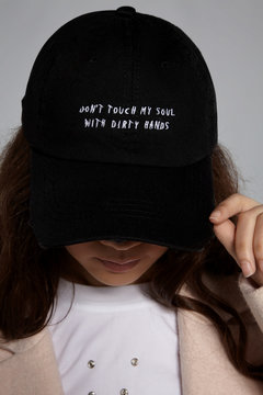 Close-up shot of brown-haired lady in a white top with steel rivets, beige jacket and a black baseball cap with an embroidered lettering "Don't touch my soul with durty hands".