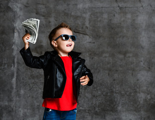 Looking up rich kid boy in sunglasses, leather jacket and red t-shirt holding a bundle of dollars...