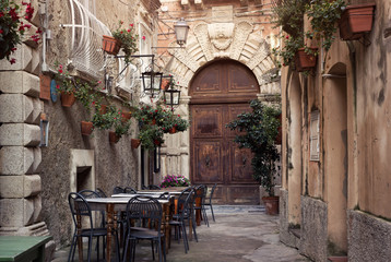 Tropea old city street with cozy restaurant terrace and floral decoration. Medieval wooden door of historical building and stone pavement.