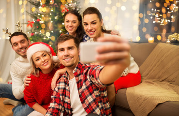 Obraz na płótnie Canvas celebration and holidays concept - happy friends with glasses celebrating christmas at home party and taking selfie by smartphone