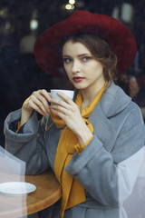 girl in a cafe with a cup of coffee and a hat. beautiful girl with a cup of coffee at the window in a cafe, waiting for a meeting, breakfast