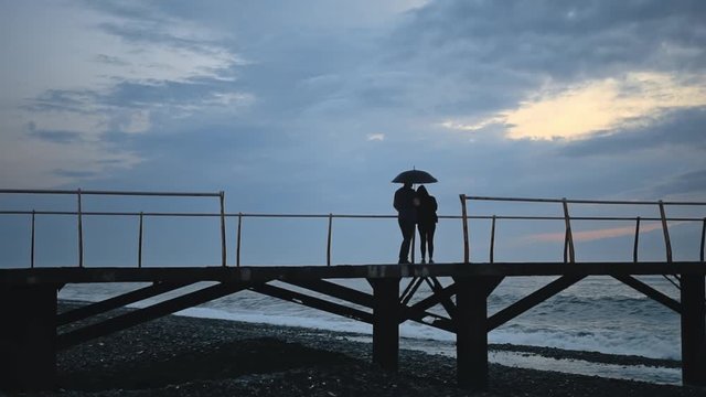 Man and girl silhouette under umbrella enjoys sunset from pier on shore of sea or ocean. Waves, storm, foam, stone beach, evening
