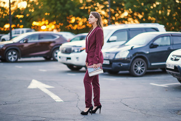 Pleasant woman in a burgundy trouser suit is standing in profile in a street car parking.