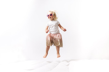 A little girl in pink sunglasses and a pink skirt jumps on a white mattress.