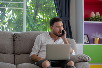 Young man is browsing at his laptop and calling with customer on the sofa at home, Business concept