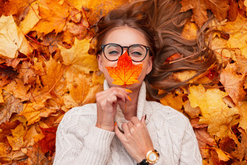 Top view on a positive young woman in stylish glasses in an knitted white sweater with an orange leaf near face in the park on a background of golden foliage. Joyful happy girl lies in the forest.
