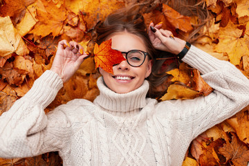 Funny young hipster woman in a fashionable knitted sweater in stylish glasses with an orange leaf in hands expresses surprise while lying on autumn foliage in the forest. Positive girl model resting.