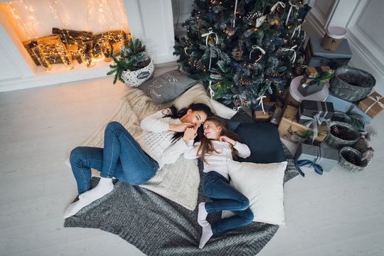 Cute kid girl and her mom playing under christmas tree with presents, lying on wooden floor in room. Wearing trendy knitted sweater, jeans. Top view.