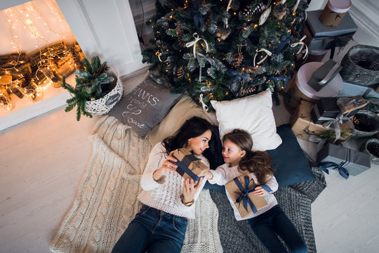 Cute kid girl and her mom playing under christmas tree with presents, lying on wooden floor in room. Wearing trendy knitted sweater, jeans. Top view.