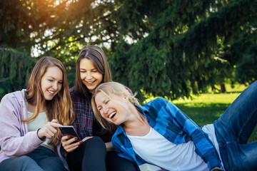 Three young girls sitting on green grass with book and smartphones. Cheerful friends resting in park. Spring and summer leisure on a nature, concept of female friendship.