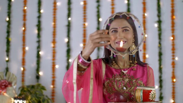 Pretty Indian lady doing Karwa Chauth Puja aginst a decorated festive background. Beautiful young woman performing Karwa Chauth rituals while looking through the sieve in traditional clothing