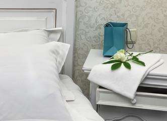 Gift package and fresh white rose on a white bedside table near a white wooden bed covered with white linens. Bouquet of flowers on a nightstand