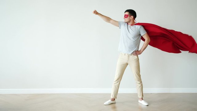 Portrait of super hero in fluttering red cape and mask standing with raised arm against empty colorful background. People, character and individuality concept.