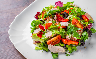 Fresh salad with chicken breast, arugula, black olives,red pepper, lettuce, fresh sald leaves and tomato on a white plate on wooden table background