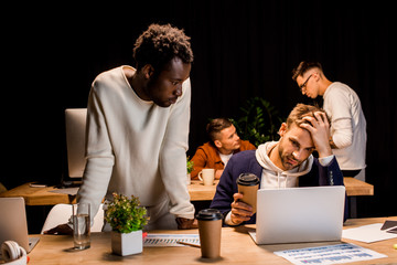african american businessman standing near exhausted colleague holding coffee to go while working at night in office
