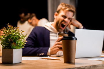 selective focus of disposable cup near exhausted businessman yawning while working at night in office