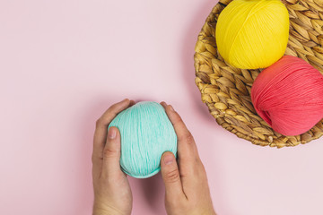 Human hands holding a blue ball of wool near to a yellow and pink balls of wool in a basket on pink background