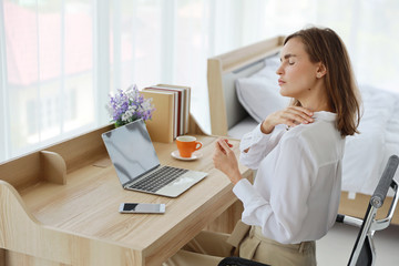 Young attractive caucasian woman wearing white casual clothes with computer and a cup of coffee. She sitting on working desk in bedroom and got pain on her shoulder with unhappy face.