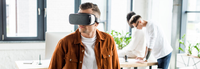panoramic shot of young businessman using vr headset while multicultural colleagues working in office