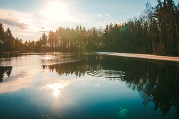 Calm morning on a forest lake. The lake is surrounded by trees. There is a little ice on the water. The sun is shining brightly