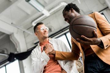 low angle view of african american businessman holding volleyball ball and shaking hands with colleague in office