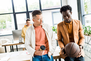 african american businessman with volleyball ball talking to colleague holding coffee to go in office