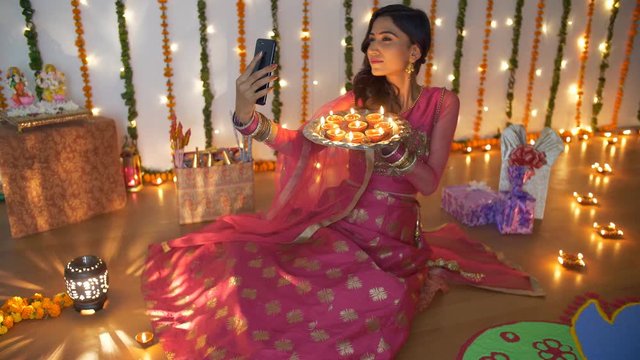Pretty Indian lady enjoying while taking selfies with burning Diyas on Diwali festival. Beautiful young female clicking pictures with her smartphone in traditional clothing - Diwali festival celebr...