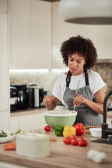 Fototapeta na wymiar Attractive mixed race woman in apron and with curly hair mixing vegetables in bowl while standing in kitchen at home.