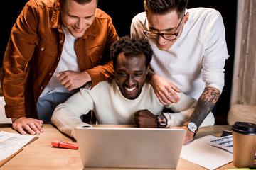 happy multicultural businessmen looking at laptop while working at night in office
