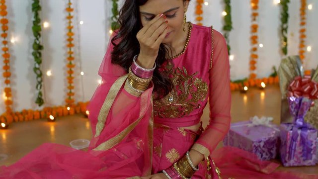 Beautiful young female smiling and putting on red bindi on her forehead on Diwali festival. Pretty Indian lady getting ready in traditional clothes on the occasion of Dipavali with decorative festi...