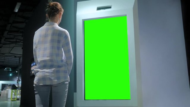 Green screen, mock up, future, copyspace, template, chroma key, technology concept. Woman looking at blank interactive touchscreen green display of electronic kiosk at futuristic exhibition or museum