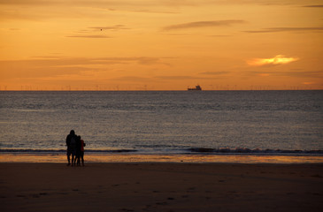 Familie macht Abendspaziergang am Meer; am Horizont scheint eine Nebensonne, fährt ein Frachter und drehen sich Windräder