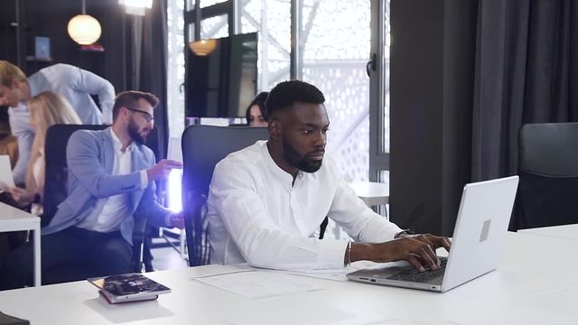 Purposeful bearded african american businessman working with papers and computer at the office table on the background of his business colleagues