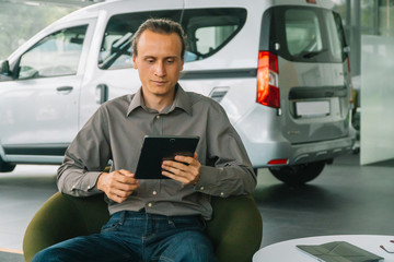 The seller in classic clothes sits in a car show on the background of a car w works with a tablet