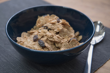 A dark blue colored bowl of delicious corn flake cereals with raisins, milk and a spoon beside a wooden surface.
