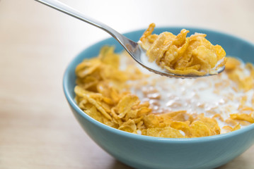 A sky blue colored bowl of delicious corn flake cereals and milk and a spoon with cereal on a wooden surface