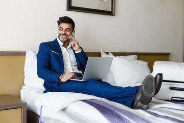 Cheerful businessman sitting on hotel bed, talking by phone