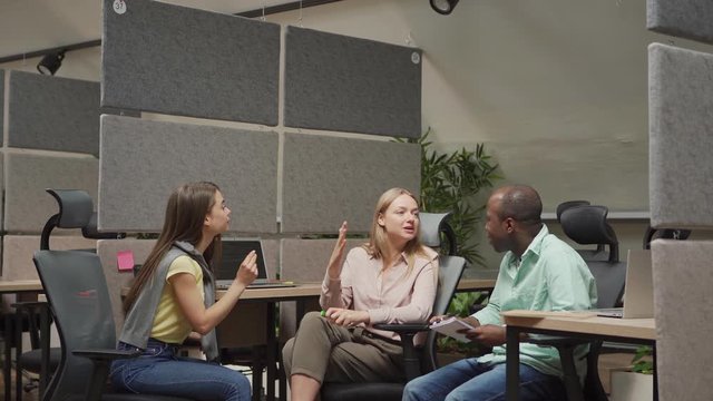 Woman in casual outfit sitting on chair and speaking with multiracial colleagues while working in modern office together