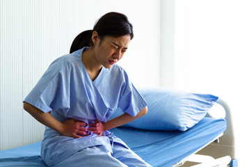 Young woman patient suffering from abdominal pain while sitting on bed at hospital. Health care...