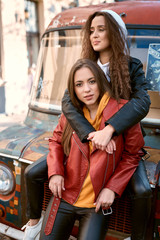 Fototapeta na wymiar Lovable caucasian women in colored leather biker jackets and hoodies posing with colored bus on background.
