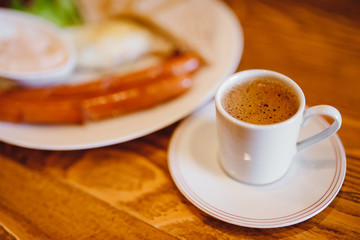 Classic English breakfast with espresso coffee in white mug, two sausages, fried eggs, toasts, lettuce on wooden background