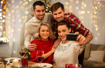 Obraz na płótnie Canvas holidays and celebration concept - happy friends taking selfie by smartphone at home christmas dinner
