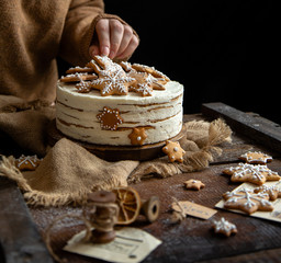 woman decorates homemade round multilayered cake with white cream and gingerbread snowflakes shaped cookies on rustic wooden table with sackcloth, fir tree branches 