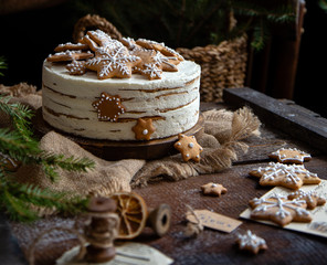 homemade round multilayered cake with white cream and gingerbread snowflakes shaped cookies on rustic wooden table with sackcloth, fir tree branches 