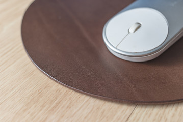 White and silver wireless mouse on a dark brown round leather mouse pad on a wooden surface.