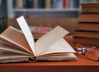 Open old book and glasses on the table, stack of old books and bookcase in the background