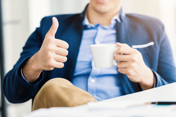 Close-up of Happy mood young businessman showing thumbs up working with holding a coffee cup sit on the table In the office room background,business expressed confidence embolden and successful